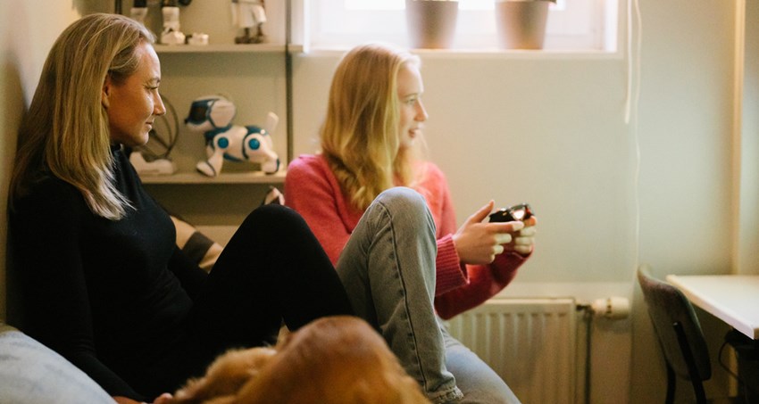 En kvinna och en flicka sitter i sen soffa i ett litet rum. De tittar på en skärm, flickan håller en handkontroll.