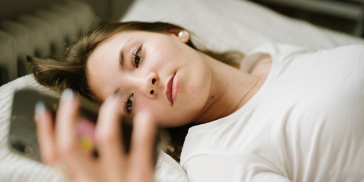 En ung kvinna ligger på rygg i en säng. Hon håller upp mobilen och tittar i den.