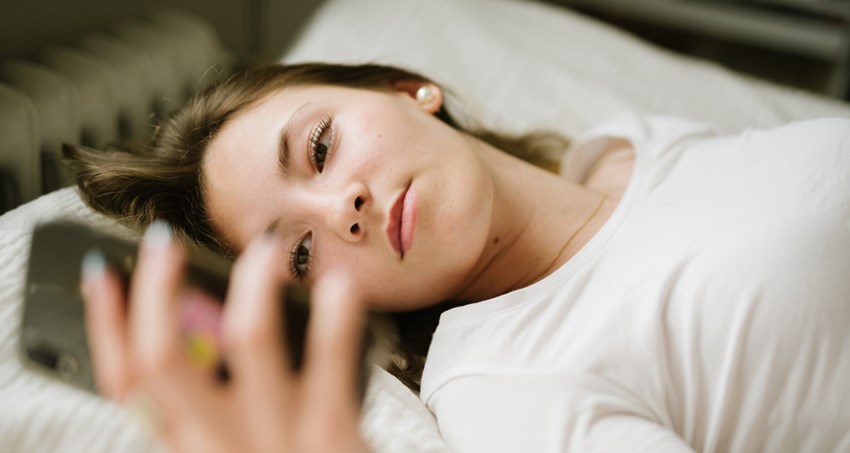 En ung kvinna ligger på rygg i en säng. Hon håller upp mobilen och tittar i den.
