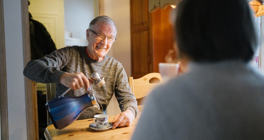 Ett äldre par sitter vid ett bord, mannen ler och häller upp en kopp kaffe