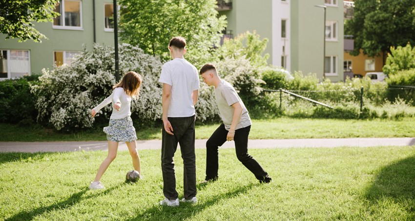 Tre tonåringar leker fotboll på en gräsmatta, i bakgrunden gångväg och hyreshus.