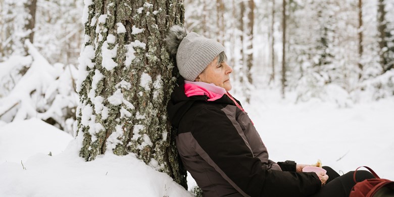 Äldre kvinna  vilar sig lutande mot ett träd i vinterlandskap
