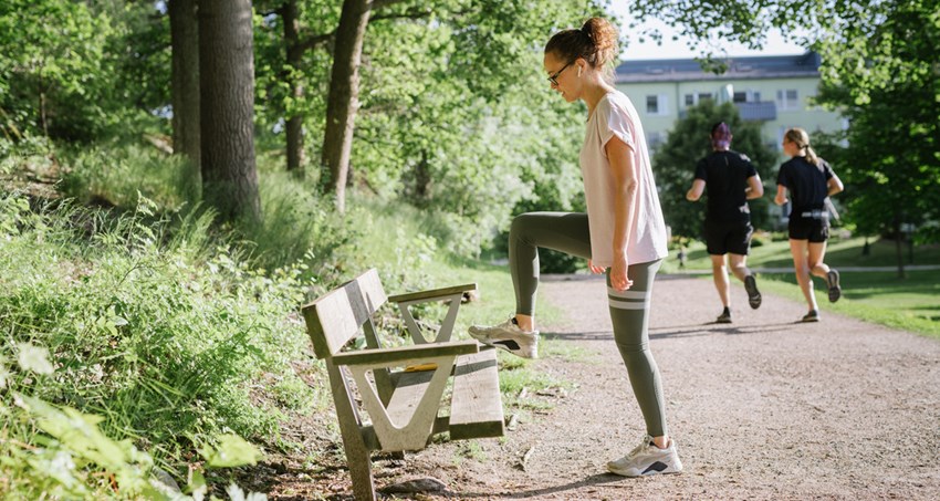 Kvinna står på en grusväg och stretchar benet mot en parkbänk.