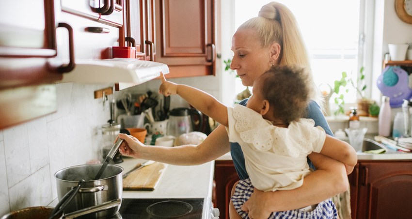 En kvinna står i ett kök och rör i en gryta på spisen. På höften bär hon ett barn.