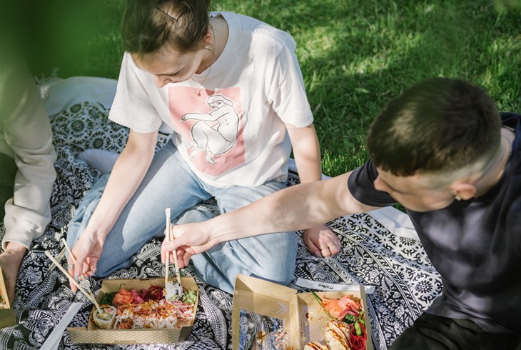 Två unga män sitter på en filt utomhus. De har varsin matlåda och äter med pinnar.