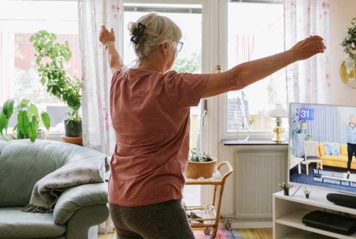Äldre kvinna gympar hemma med instruktör på TVn.