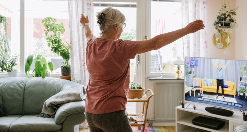 Äldre kvinna gympar hemma med instruktör på TVn.