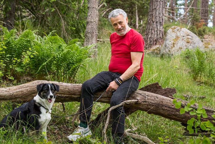 En man sitter på en stock i skogen . han har en hund i koppel.
