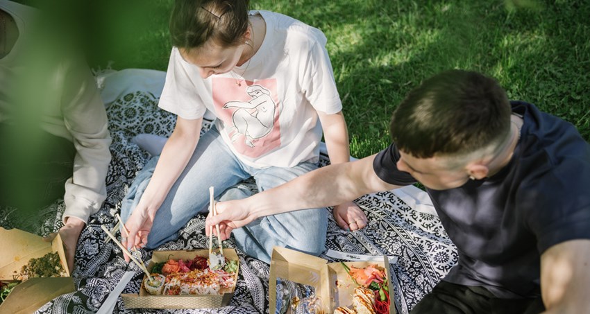 Två unga män sitter på en filt utomhus. De har varsin matlåda och äter med pinnar.
