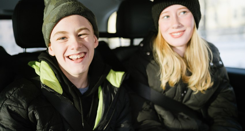 En glad kille och en glad tjej sitter med ytterkläderna på i baksätet på en bil.