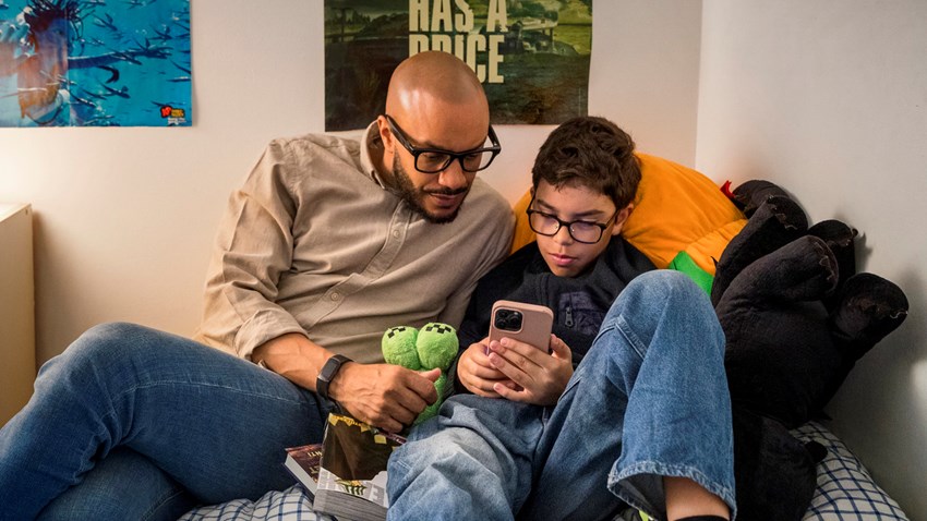 En man och ett barn sitter i  en säng och tittar på barnets mobil