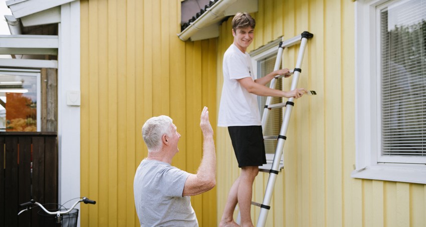 En äldre man pratar med en yngre som står på en stege lutad mot en husvägg.
