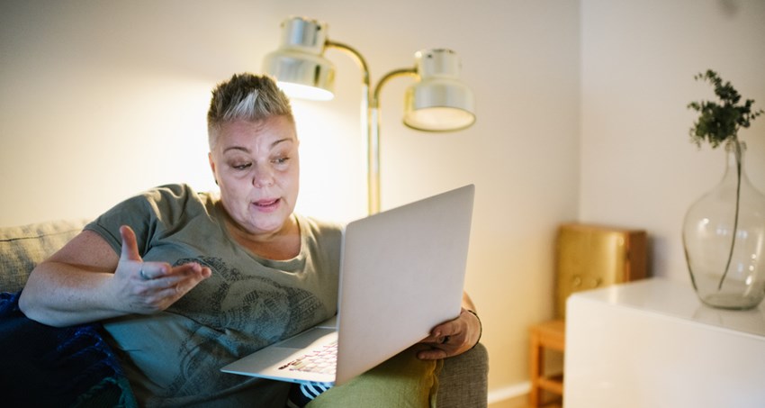 En kvinna sitter i en soffa, hon tittar i en öppen laptop som står på armstödet.