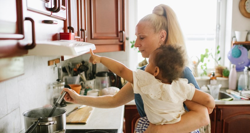 En kvinna står i ett kök och rör i en gryta på spisen. På höften bär hon ett barn.
